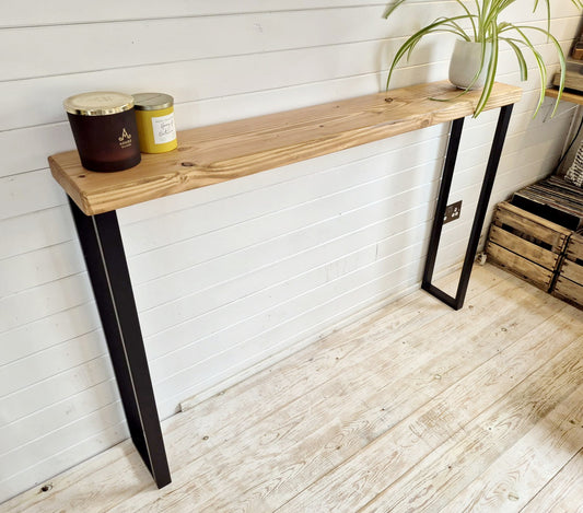 A narrow console table with sleek square legs made of dark material, paired with a light wood tabletop, placed against a white wall with a plant and decorative items on top.